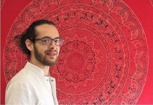Homme souriant devant un mandala rouge, portant des lunettes. D'Ici et de Claire